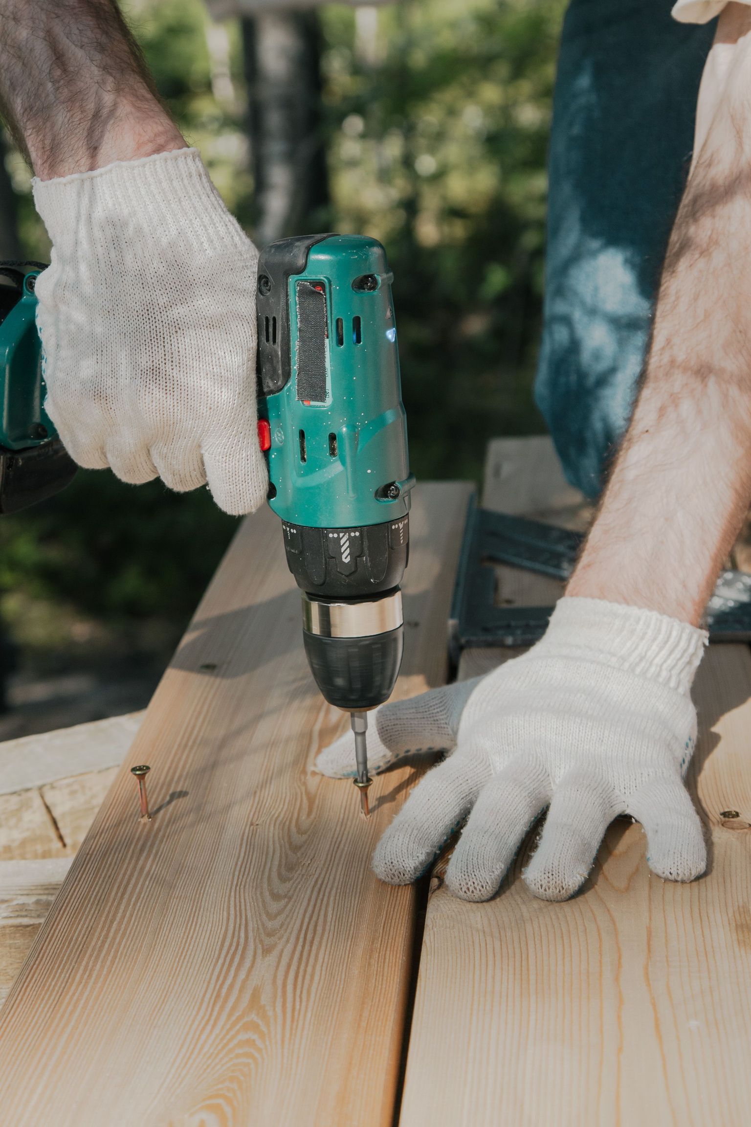Adult using a power drill on a wooden surface, wearing white gloves, outdoors in daylight.