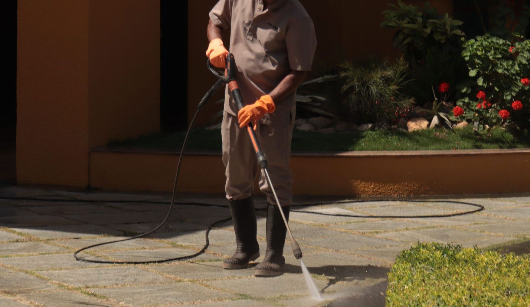 A gardener wearing a hat cleans an urban sidewalk with a pressure washer.